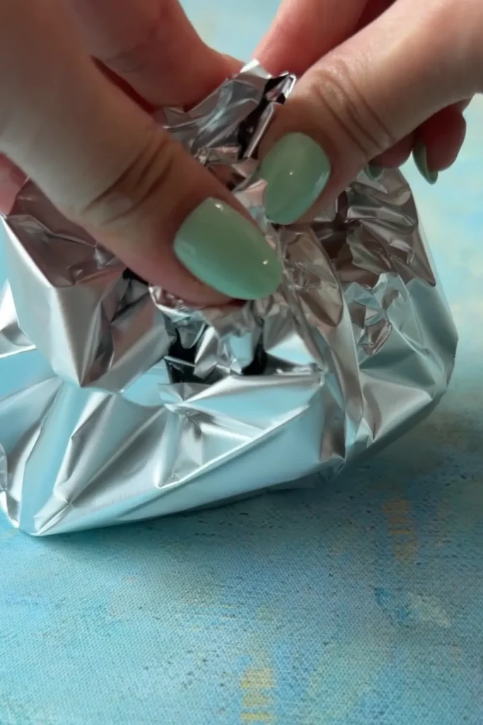 Hands wrapping a seasoned garlic head in foil, folding the foil up and around it to seal it before cooking.