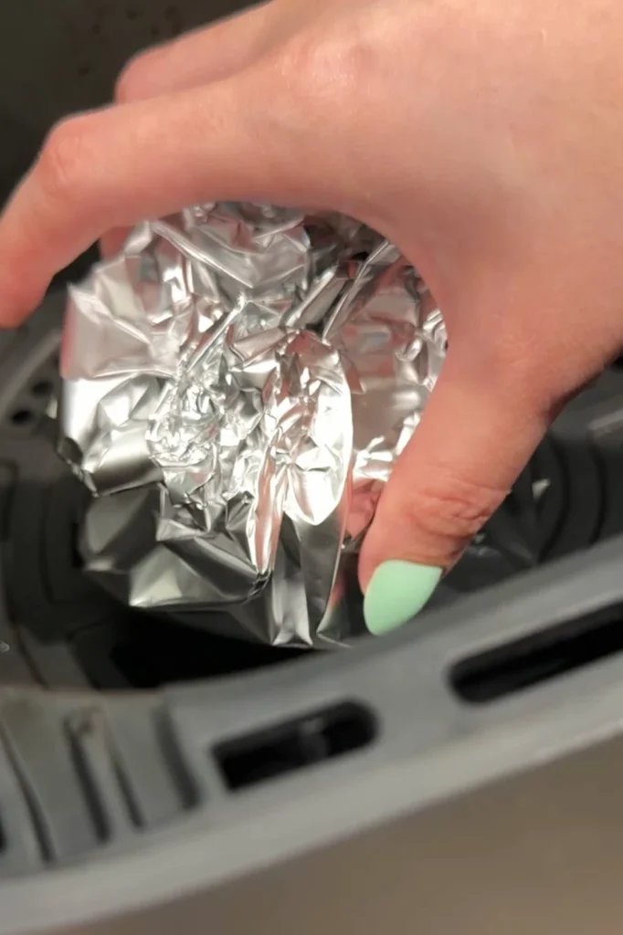A foil-wrapped head of garlic being placed into the basket of an air fryer by hand, ready to roast.
