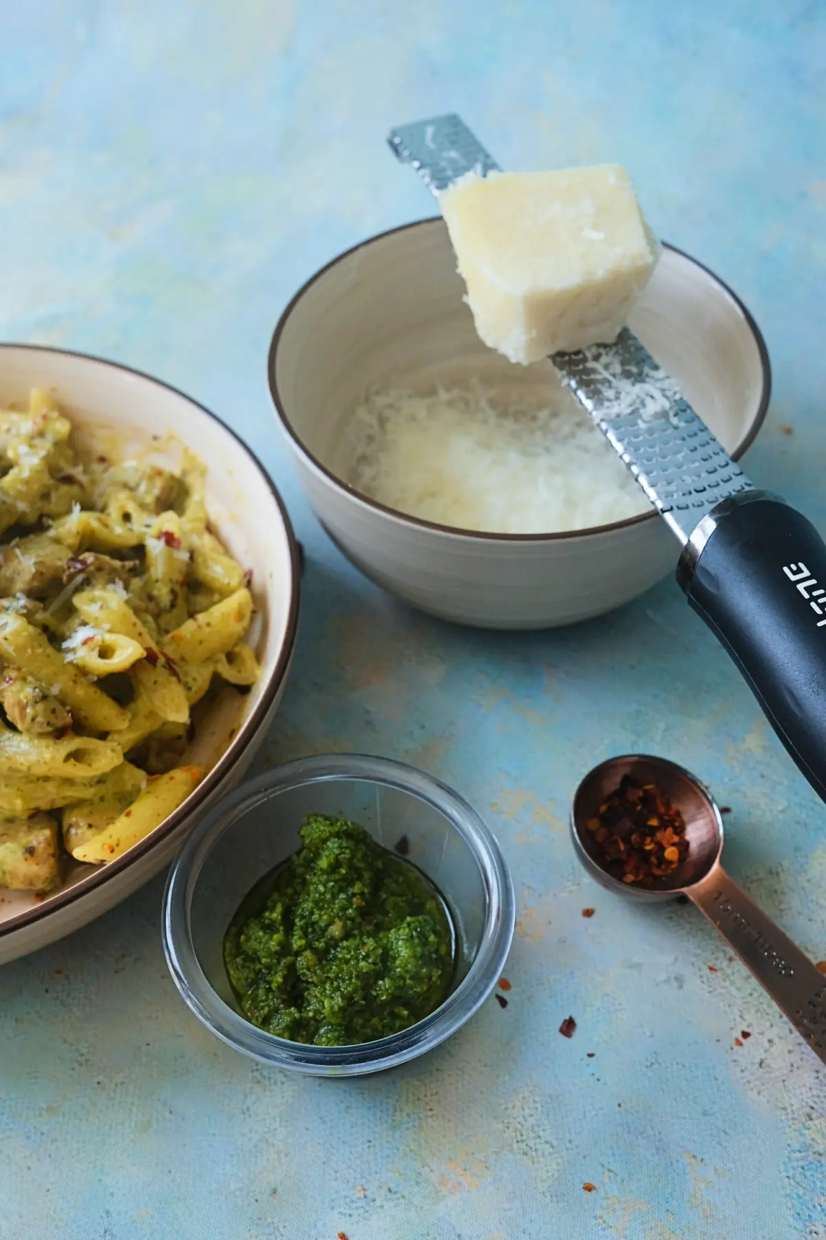 Freshly grated parmesan on a microplane over a small bowl, with almond pesto and chili flakes nearby, showing prep elements for pesto pasta.