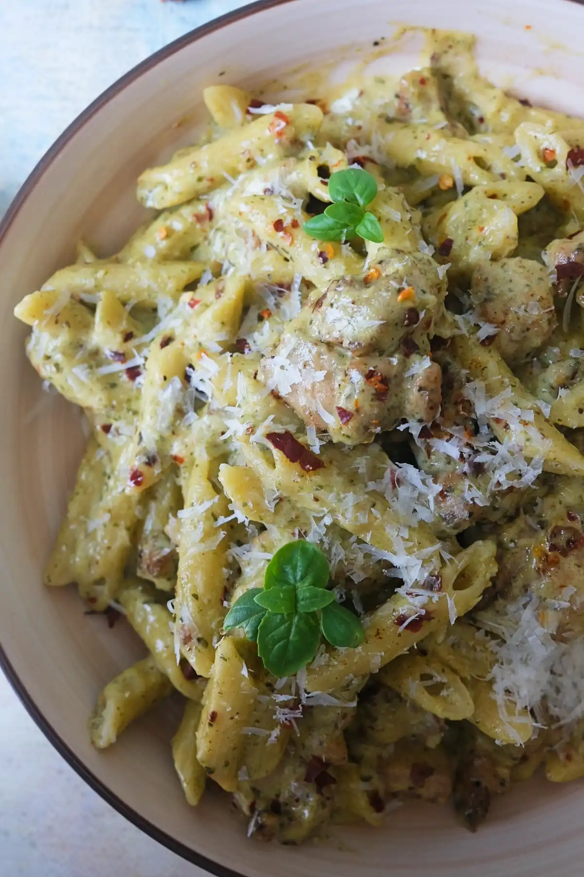 Creamy almond pesto pasta with penne and chicken, coated in a rich green basil sauce, topped with grated parmesan, fresh basil, and red pepper flakes in a shallow bowl.