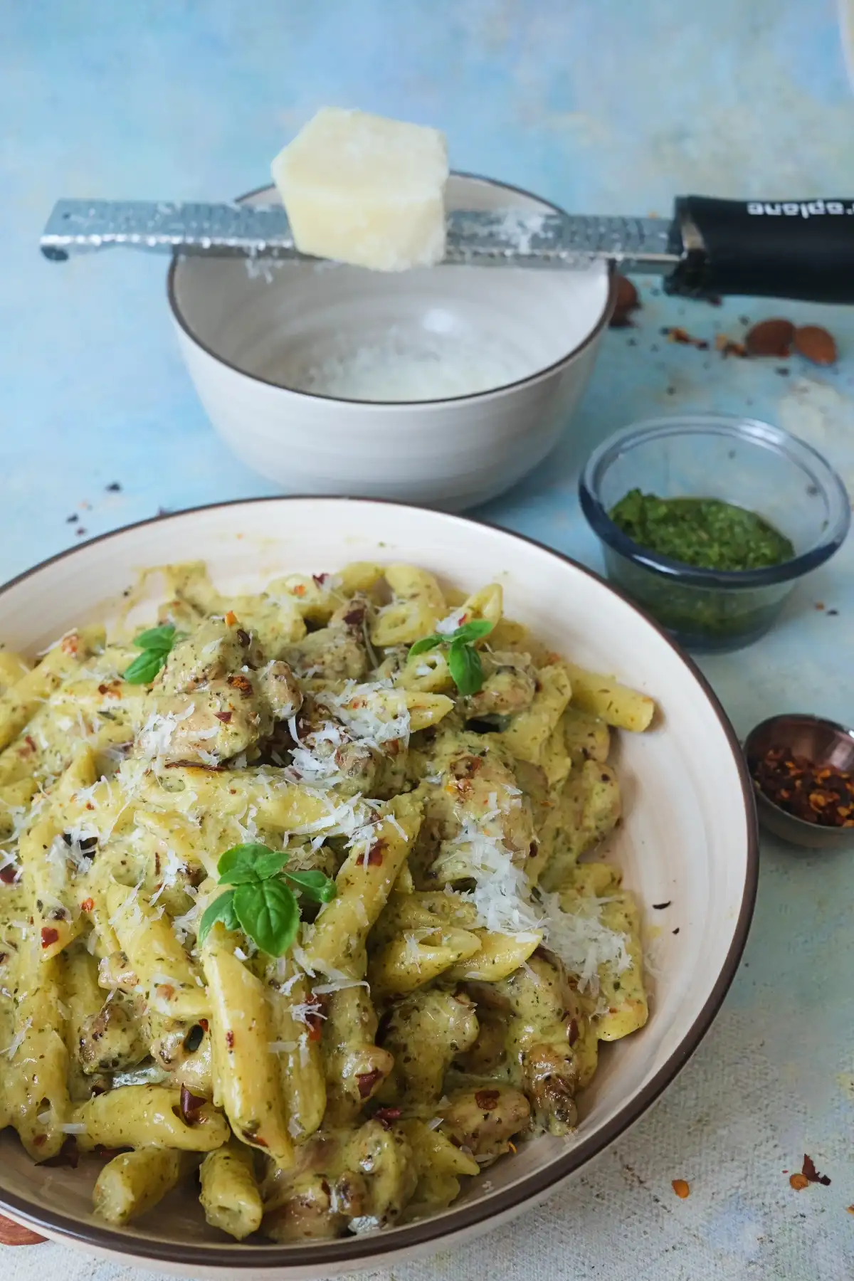 Creamy almond pesto pasta with chicken, parmesan, and basil garnish in a shallow bowl, styled with pesto and chili flakes on a blue background.