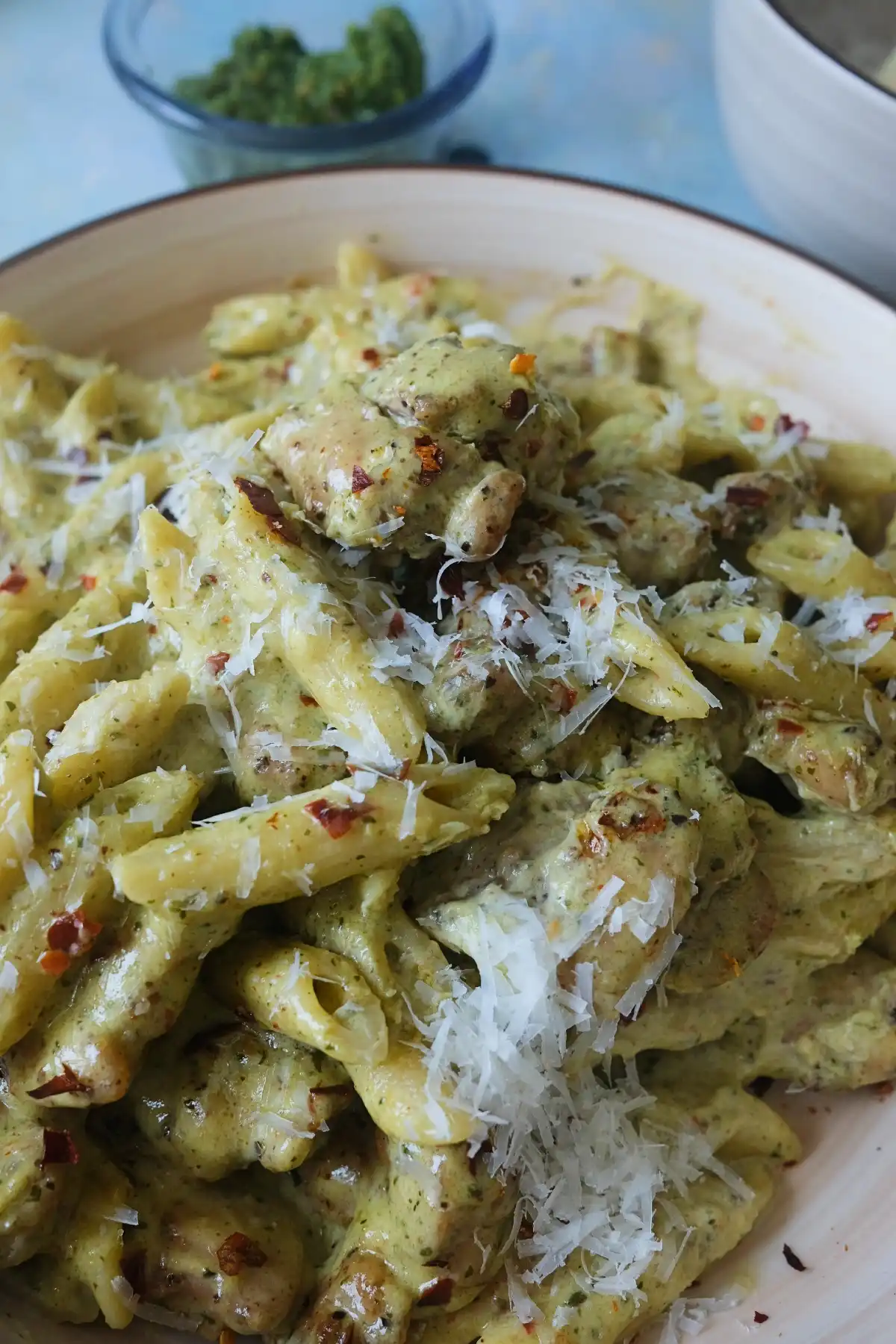 Close-up of almond pesto chicken pasta with penne, creamy basil sauce, parmesan shavings, and chili flakes, showing the thick, glossy texture of the sauce.