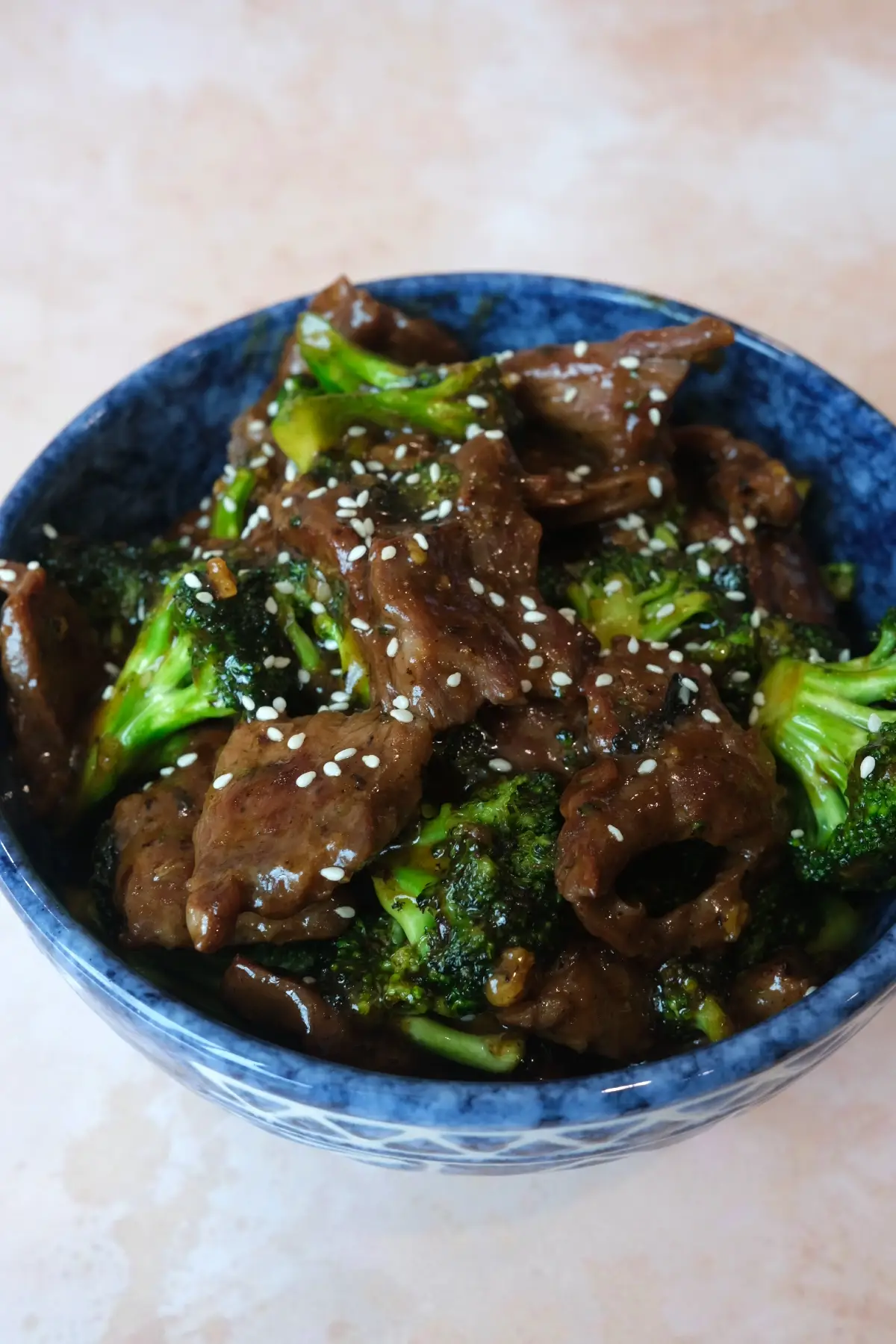 Blue bowl filled with saucy beef and broccoli, topped with white sesame seeds, shown at a slightly angled close-up on a light pink surface.