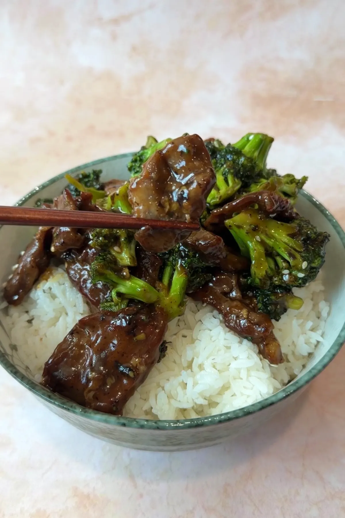 Chopsticks holding a glossy piece of beef above a bowl of beef and broccoli over white rice, with bright green broccoli and saucy sliced beef underneath.
