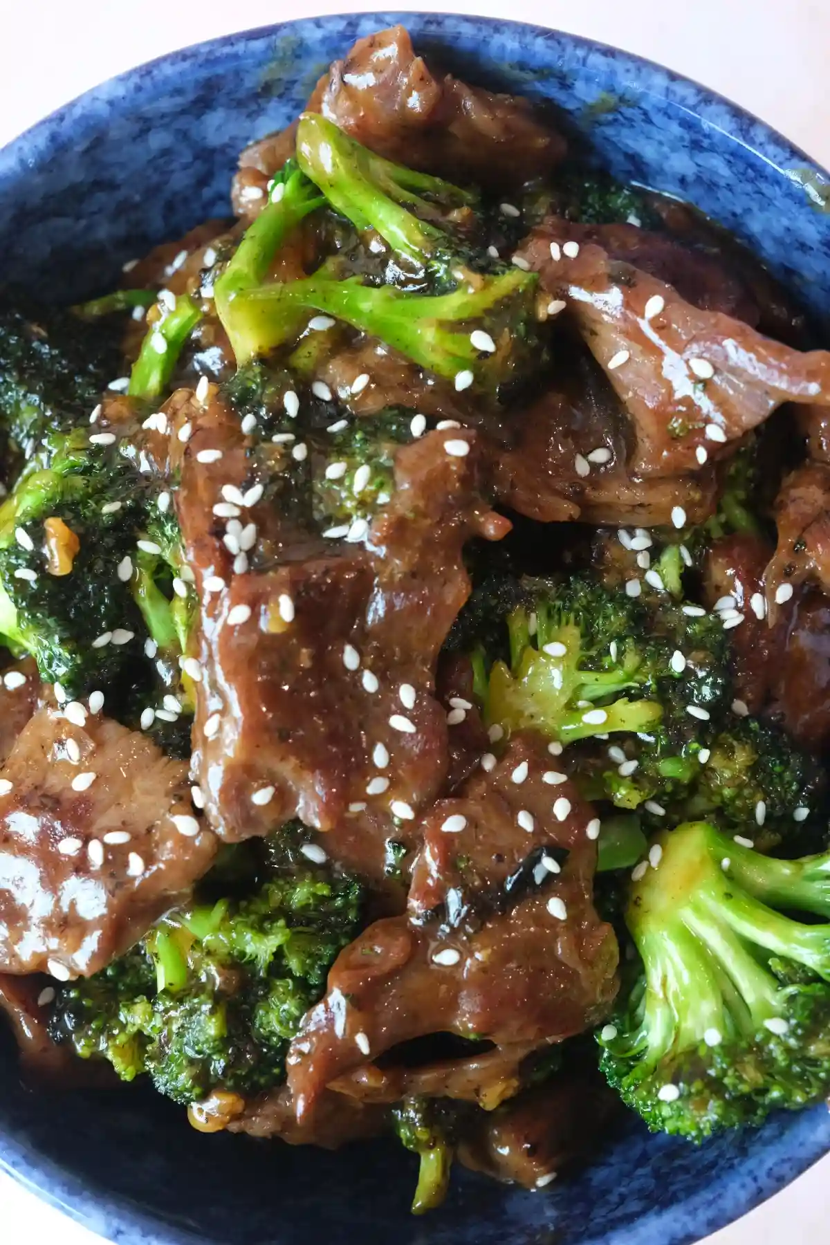 Close-up of Panda Express beef and broccoli in a blue ceramic bowl, with glossy sliced beef, bright green broccoli, and white sesame seeds on top.