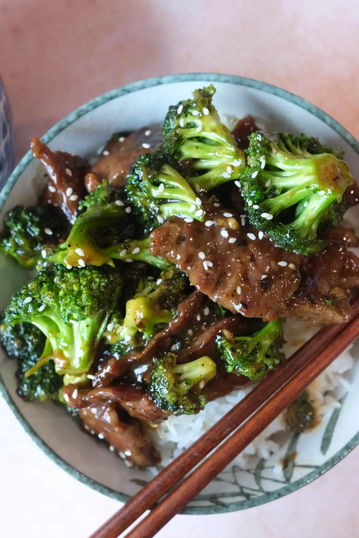 Beef and broccoli served over white rice in a patterned bowl with chopsticks on the side, topped with sesame seeds and glossy brown sauce.