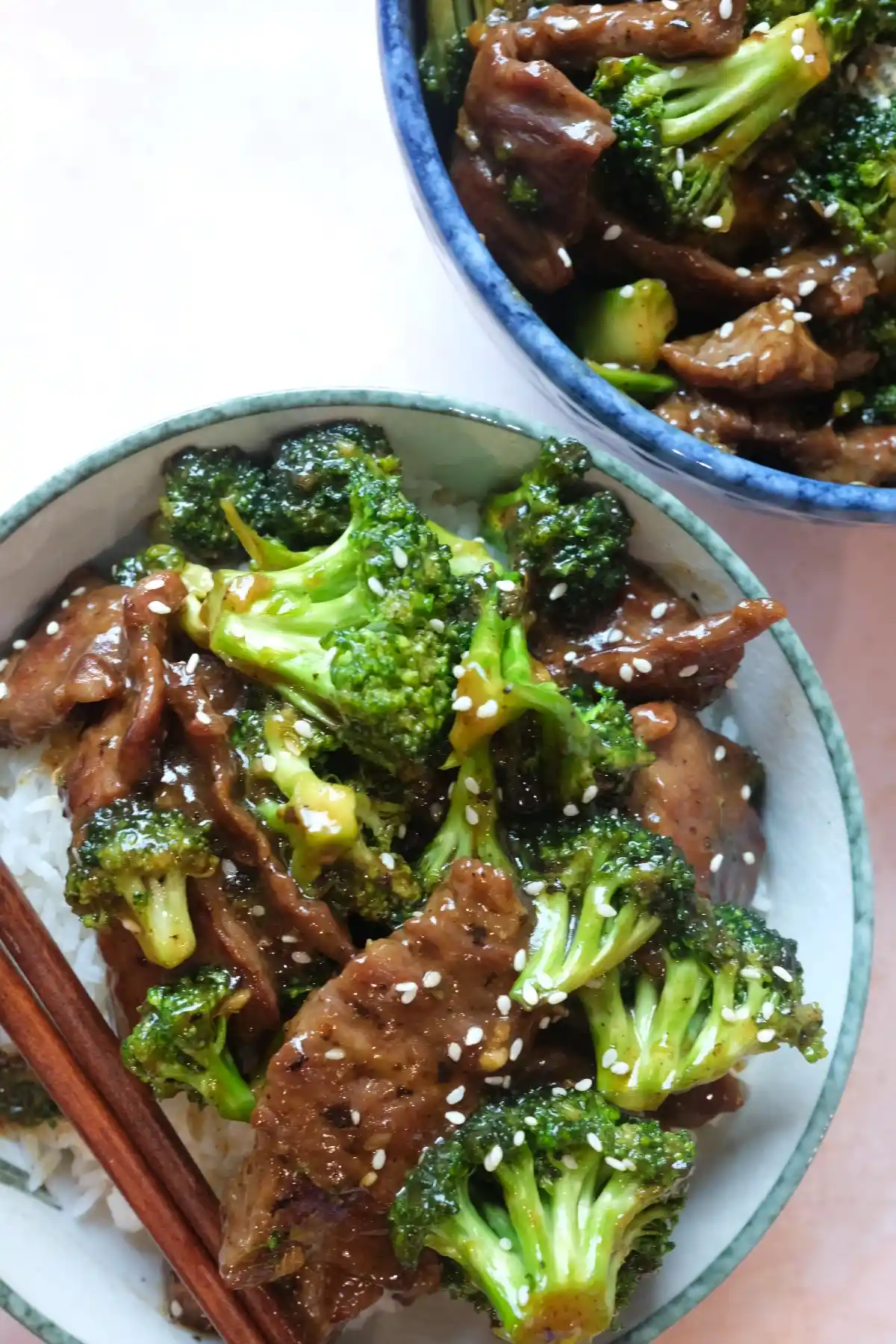 Two bowls of beef and broccoli over rice, with a close-up rice bowl in front and a larger blue serving bowl in the background, both topped with sesame seeds.
