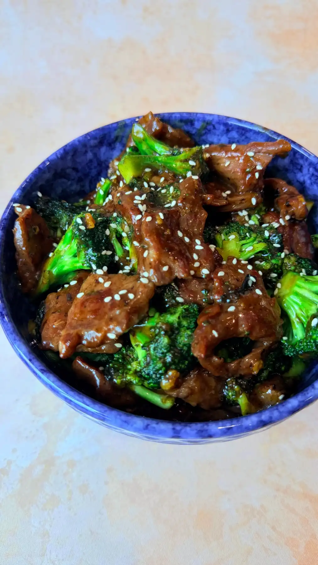 Blue patterned bowl filled with glossy beef and broccoli, garnished with sesame seeds and photographed on a soft pink background with extra space above.