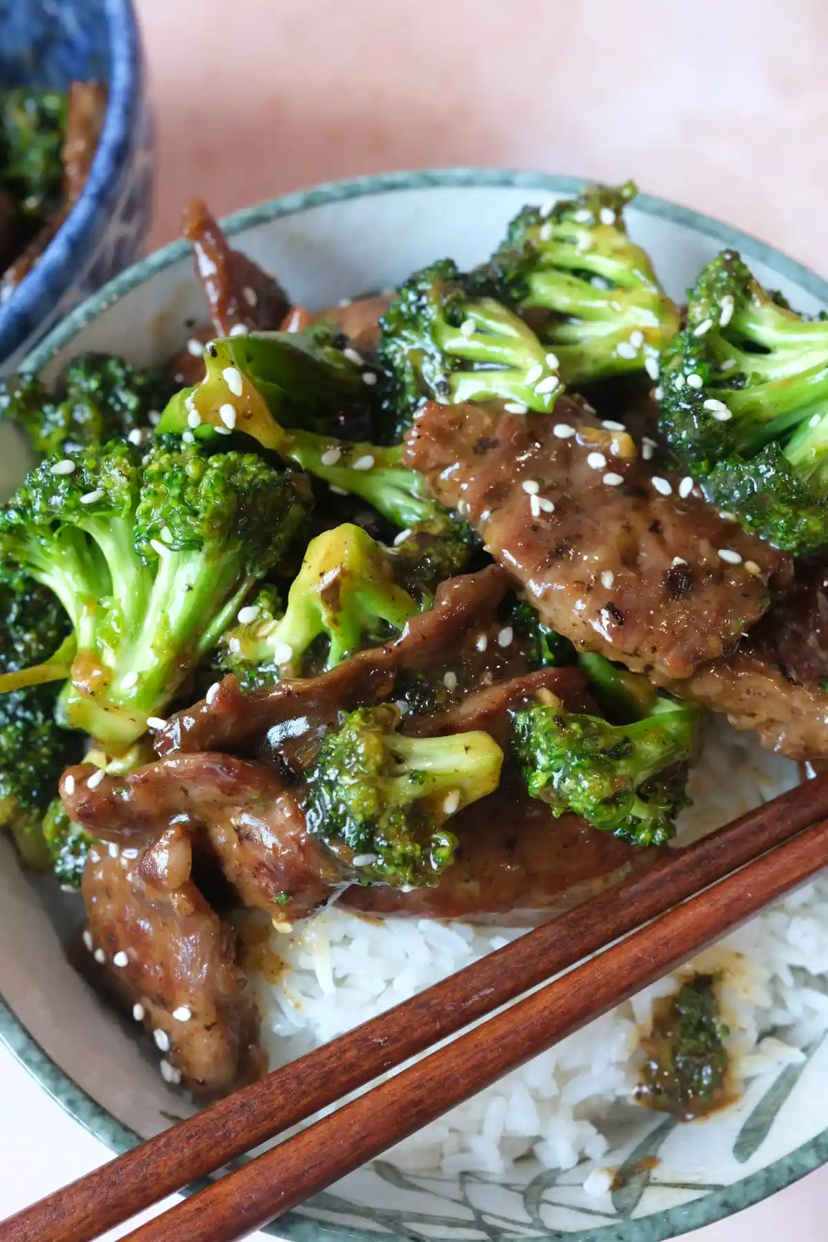 Bowl of beef and broccoli over white rice with wooden chopsticks across the front, with a second blue bowl of beef and broccoli blurred in the background.