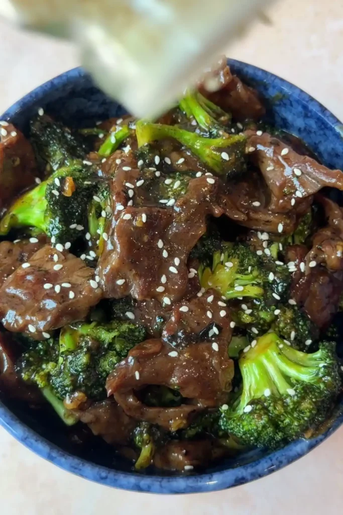 White sesame seeds being sprinkled over the finished beef and broccoli in a blue serving bowl.