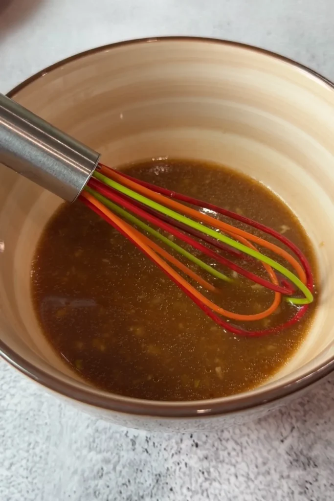 Whisk resting in a beige bowl of mixed beef and broccoli sauce with garlic and ginger on a light countertop.