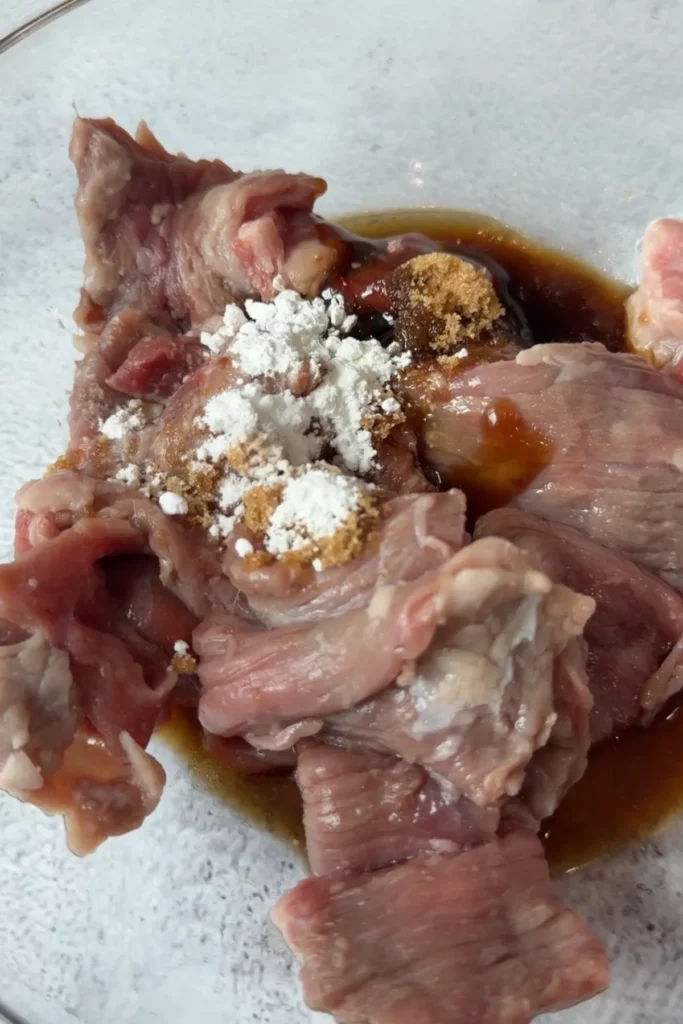 Thinly sliced raw beef in a glass bowl with soy sauce, oyster sauce, brown sugar, and cornstarch added on top for the marinade.