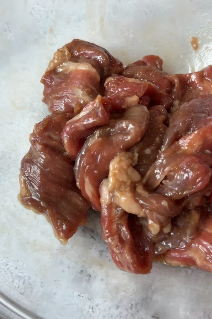 Sliced beef fully coated in a glossy brown marinade in a glass mixing bowl.