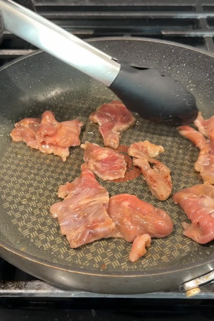 Marinated beef slices added to a hot skillet in a single layer with silicone-tipped tongs.