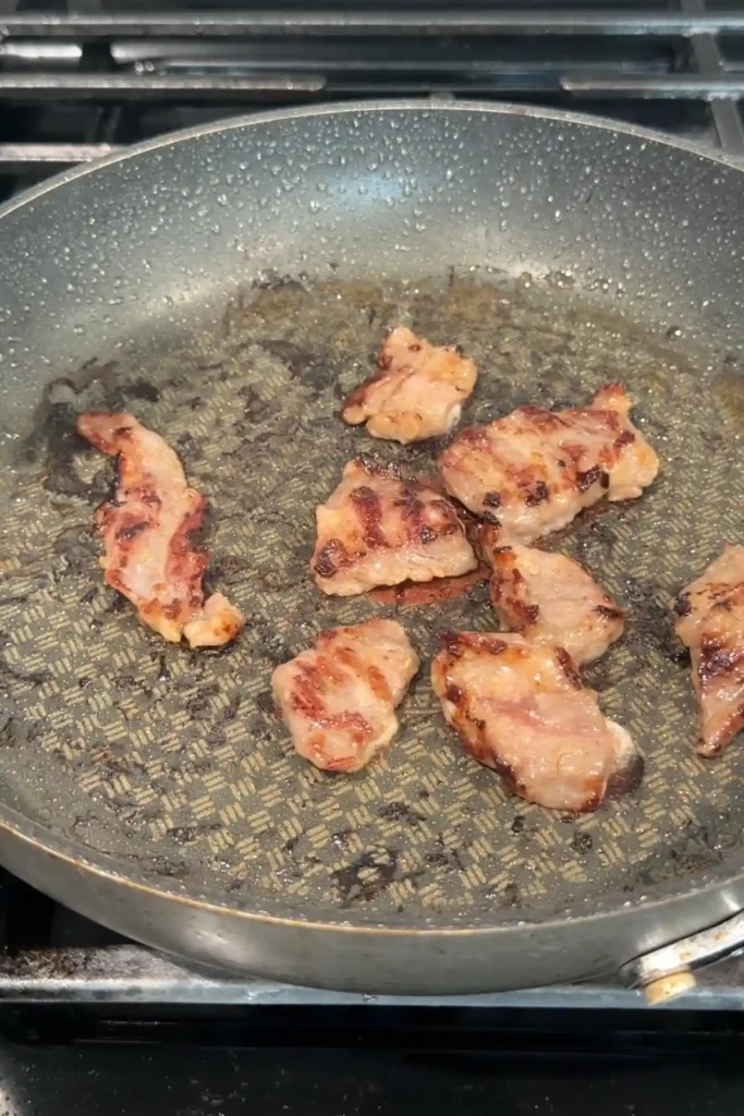 Thin slices of beef browning in a skillet with seared edges after being cooked on both sides.