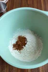 Dry ingredients for carrot cupcakes in a large light green mixing bowl, with flour and ground cinnamon piled together before mixing on a wooden countertop.