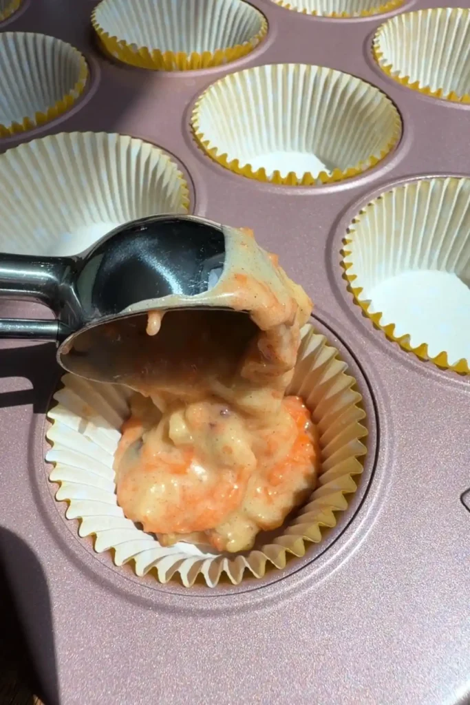 A cookie scoop dropping carrot cupcake batter into a paper liner in a muffin pan for evenly sized cupcakes.
