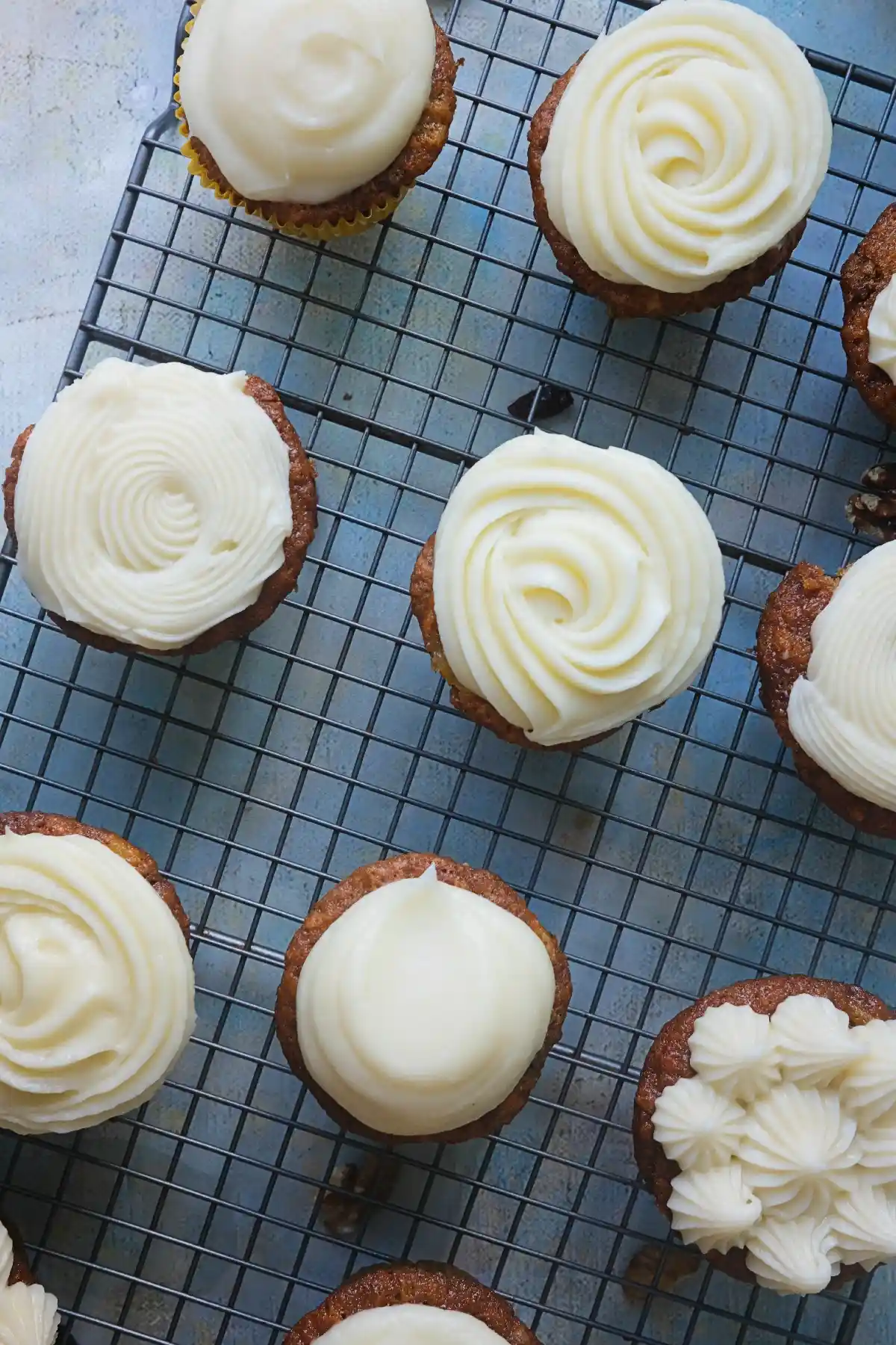 Overhead shot of carrot cake cupcakes on a cooling rack, each topped with cream cheese frosting in different decorative patterns such as spirals, smooth dollops, and piped stars.