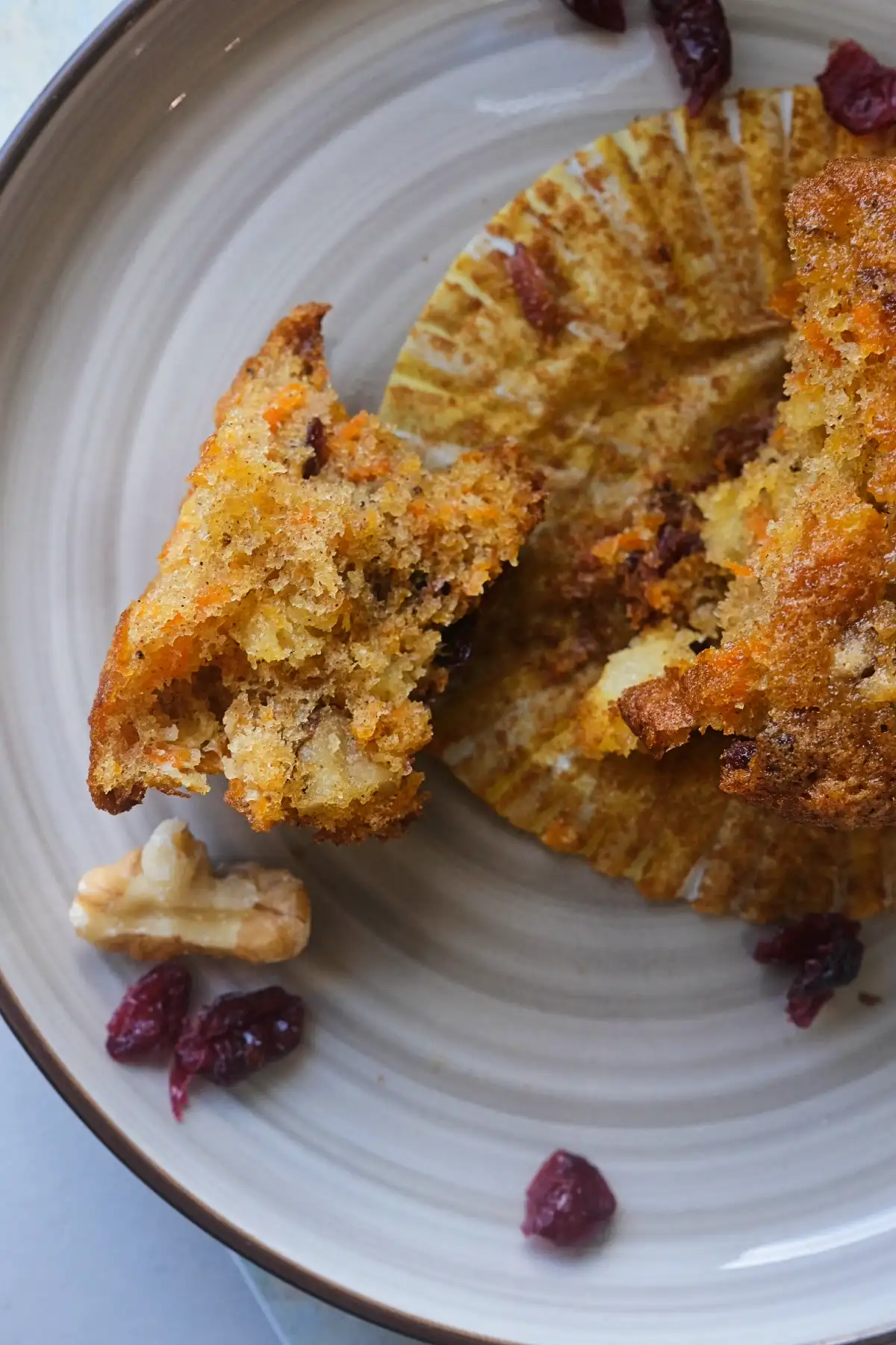 Close-up of a carrot cake cupcake torn in half on a gray plate, showing a moist crumb studded with shredded carrot, chopped walnuts, crushed pineapple, and dried cranberries.