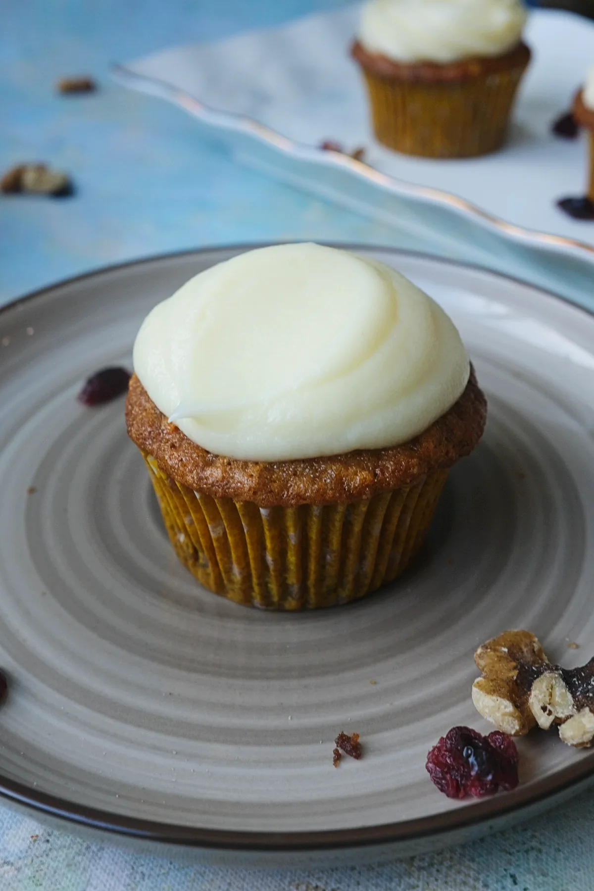 Single carrot cake cupcake on a gray plate topped with a smooth dome of cream cheese frosting, with a white serving platter and more frosted cupcakes blurred in the background.