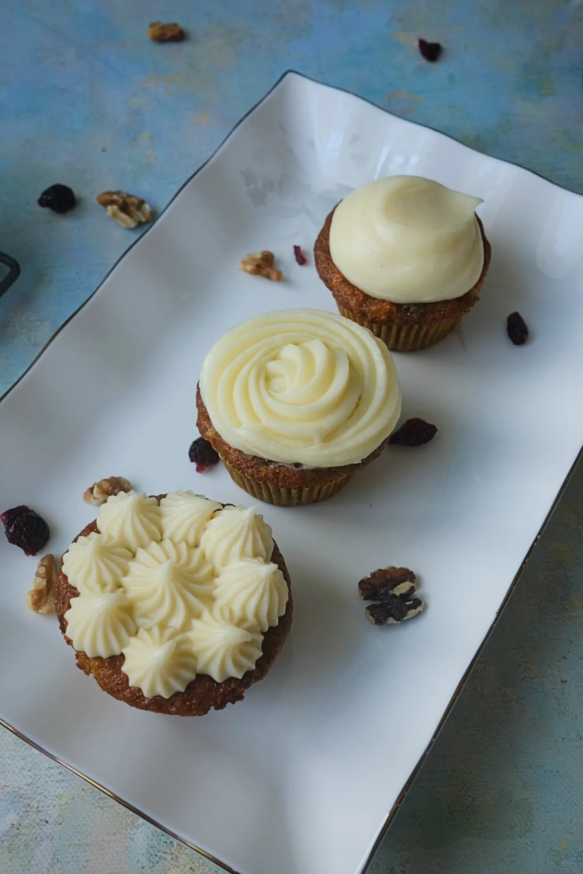 Three carrot cake cupcakes on a white rectangular platter, each finished with a different cream cheese frosting design , with walnuts and dried cranberries scattered around.