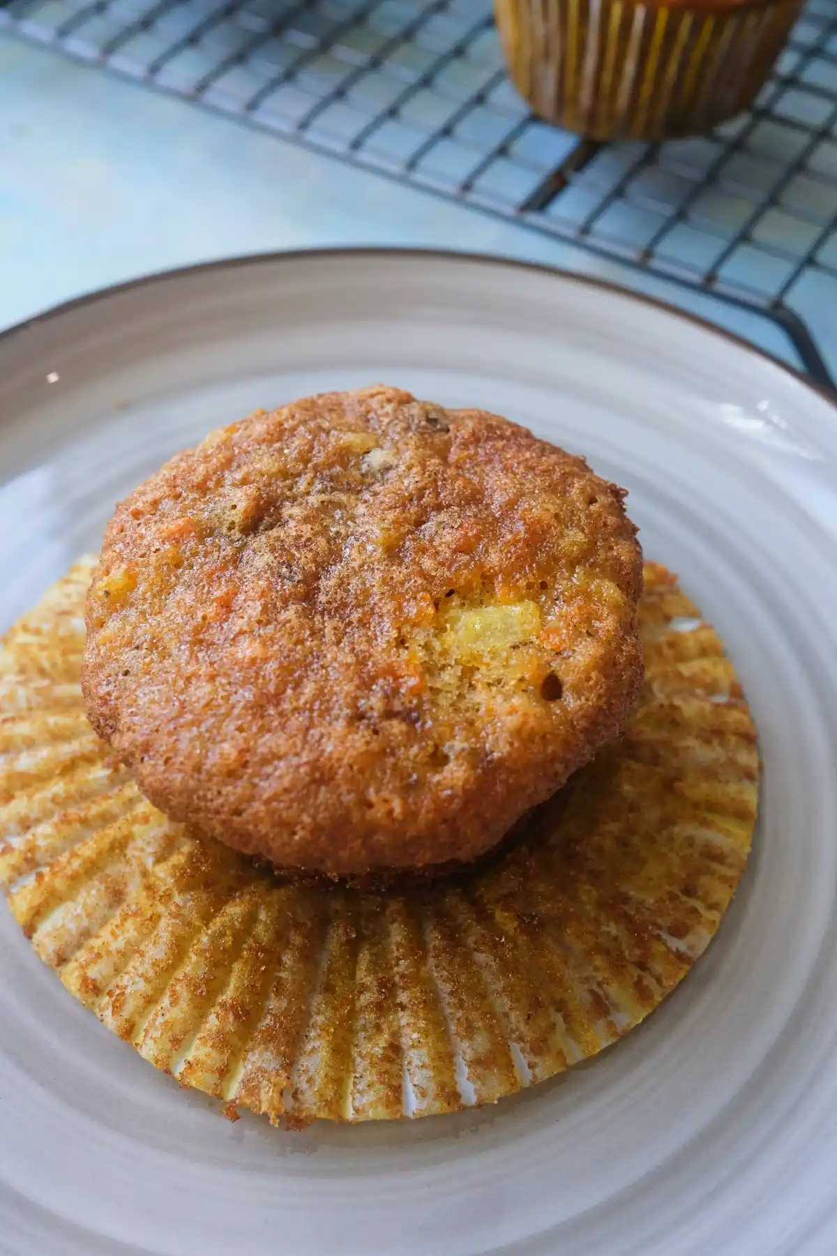Plain carrot cake cupcake without frosting resting on its opened paper liner on a gray plate, showing a golden brown top with visible bits of carrot and pineapple.