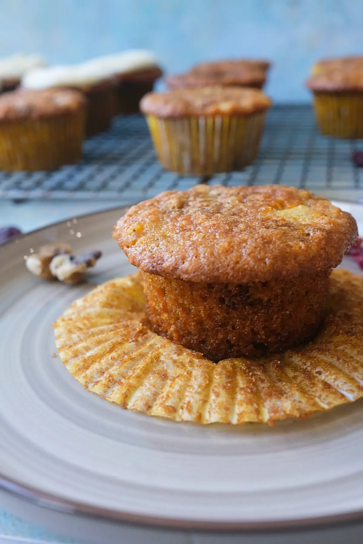 Side view of an unfrosted carrot cake cupcake on its flattened liner, highlighting the tall cupcake shape, moist golden crumb, and slightly domed top, with more cupcakes on a rack in the background.