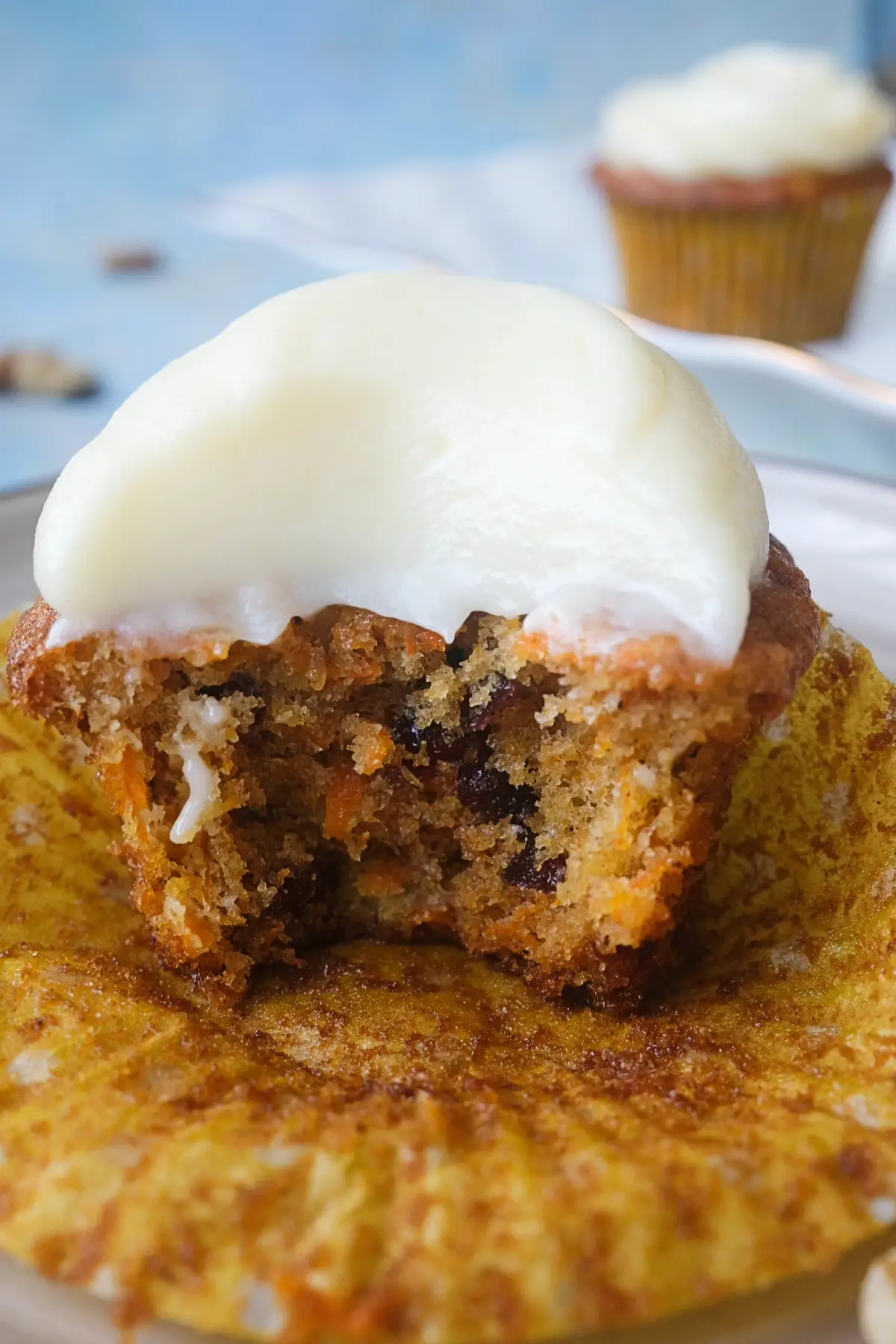 Close-up of a carrot cake cupcake with a large bite taken out, topped with a thick layer of cream cheese frosting and revealing a moist interior filled with carrot shreds, dried cranberries, walnuts, and pineapple.
