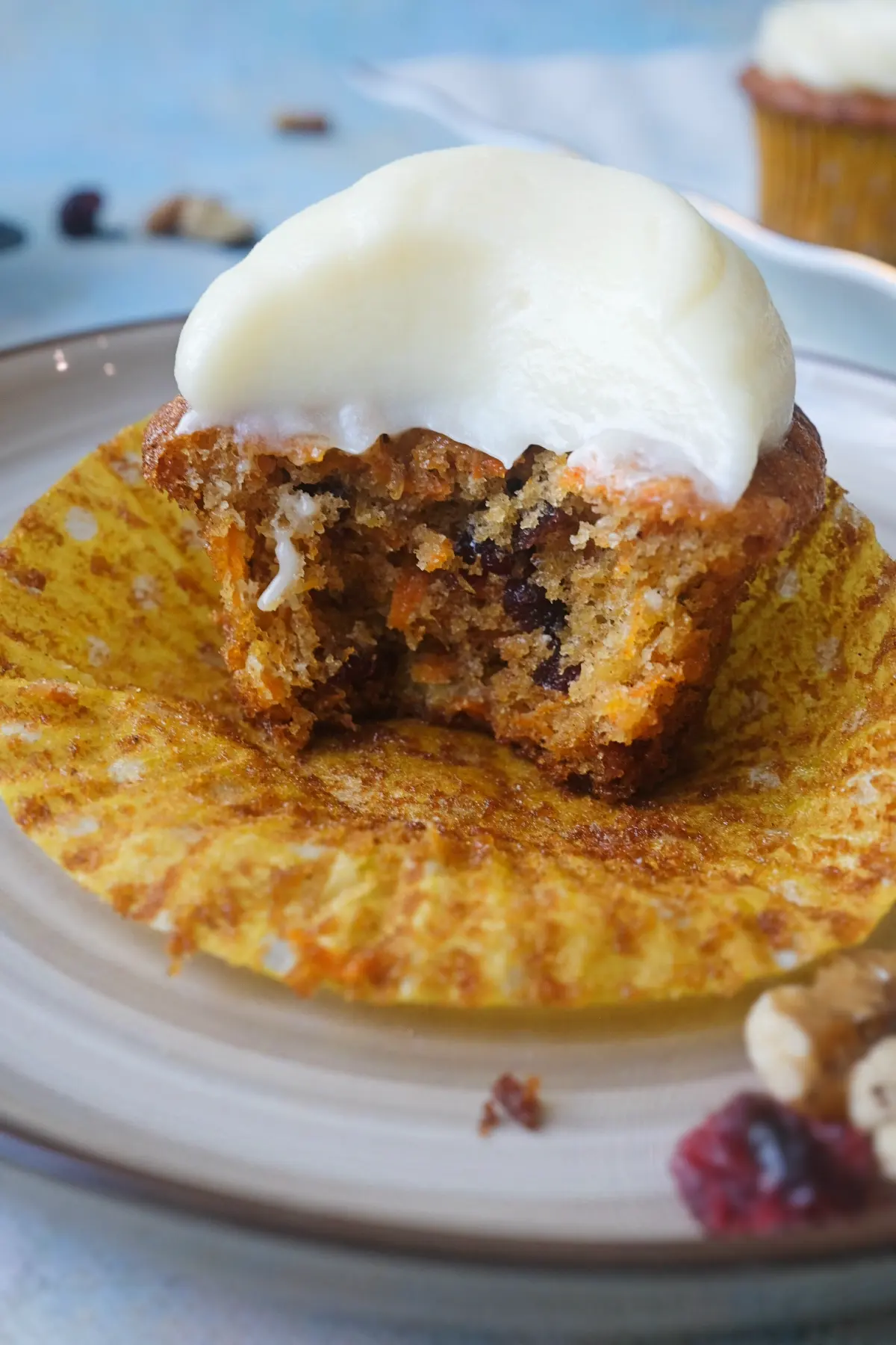 Close-up of a bitten carrot cake cupcake on its opened liner with a thick cap of cream cheese frosting, showing the soft crumb packed with grated carrot, dried cranberries, walnuts, and small bits of pineapple.