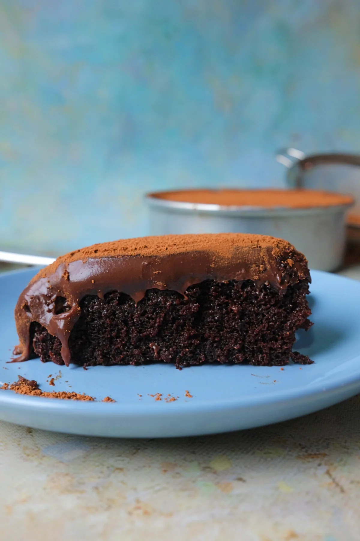 Slice of chocolate dream cake on a blue plate, showing a moist, dark chocolate crumb with a thick layer of glossy ganache dripping down the sides, finished with a dusting of cocoa powder on top.
