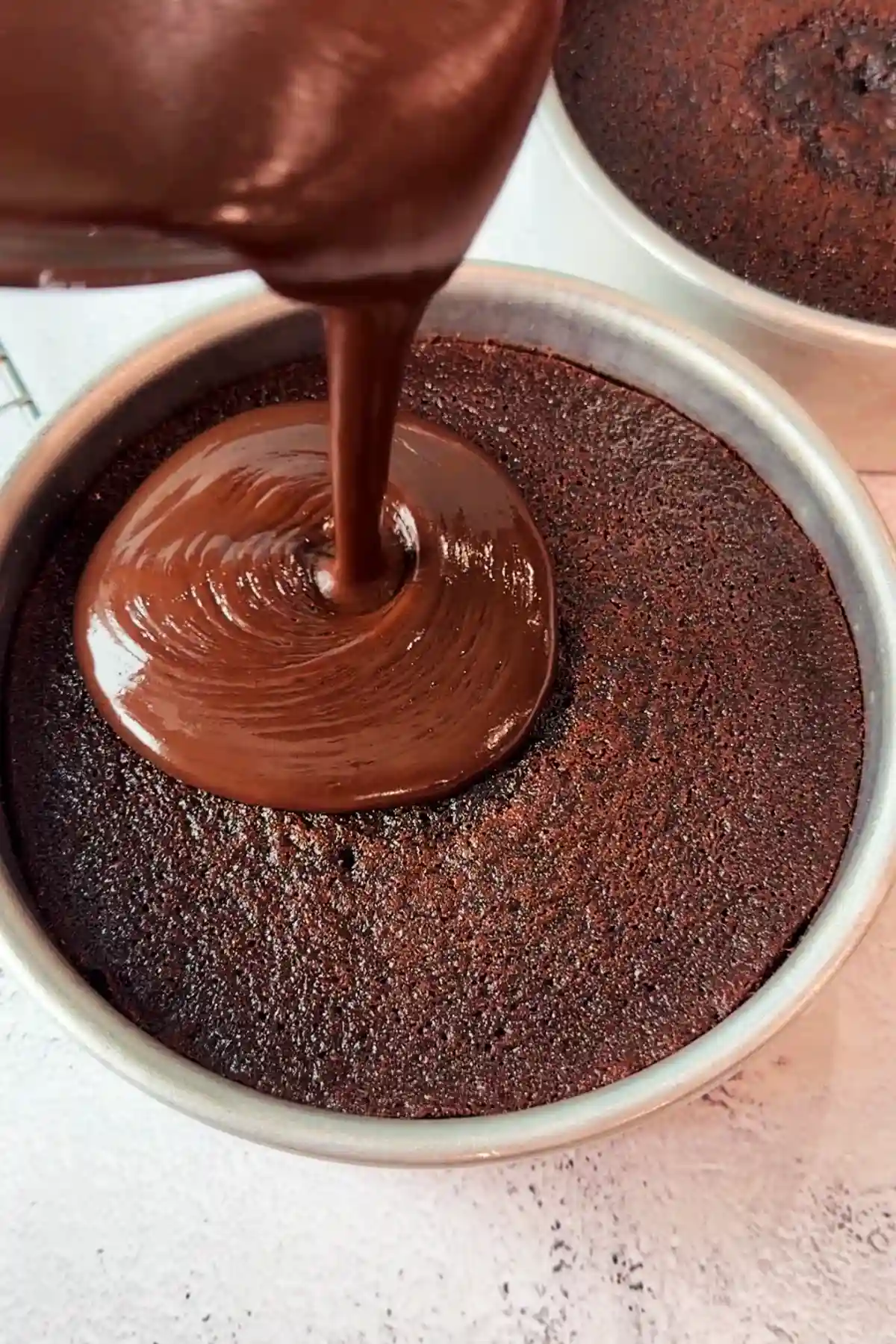 Ganache being poured onto a baked cake layer in a pan, spreading outward from the center in a thick, even layer.