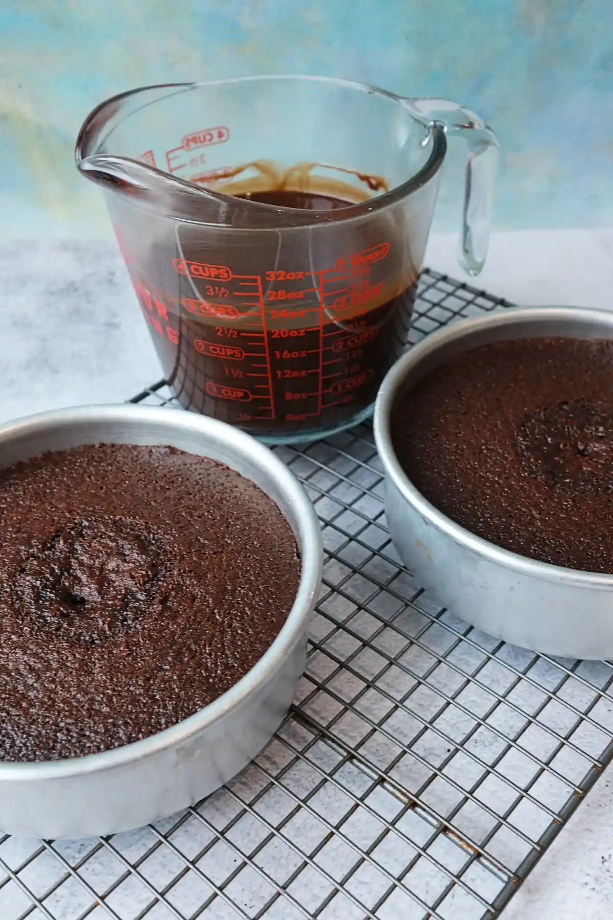 Two freshly baked chocolate cake layers in round metal pans cooling on a wire rack, with a glass measuring cup of smooth, melted chocolate ganache behind them against a light blue background.