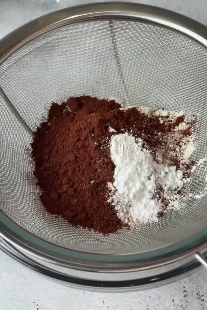 Dry ingredients including cocoa powder, flour, and leavening agents sifted together in a fine mesh strainer over a bowl.