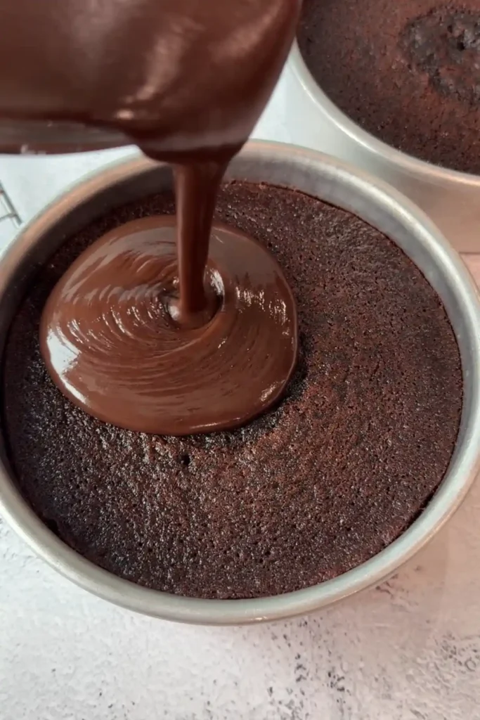 Ganache being poured onto a baked cake layer in a pan, spreading outward from the center in a thick, even layer.