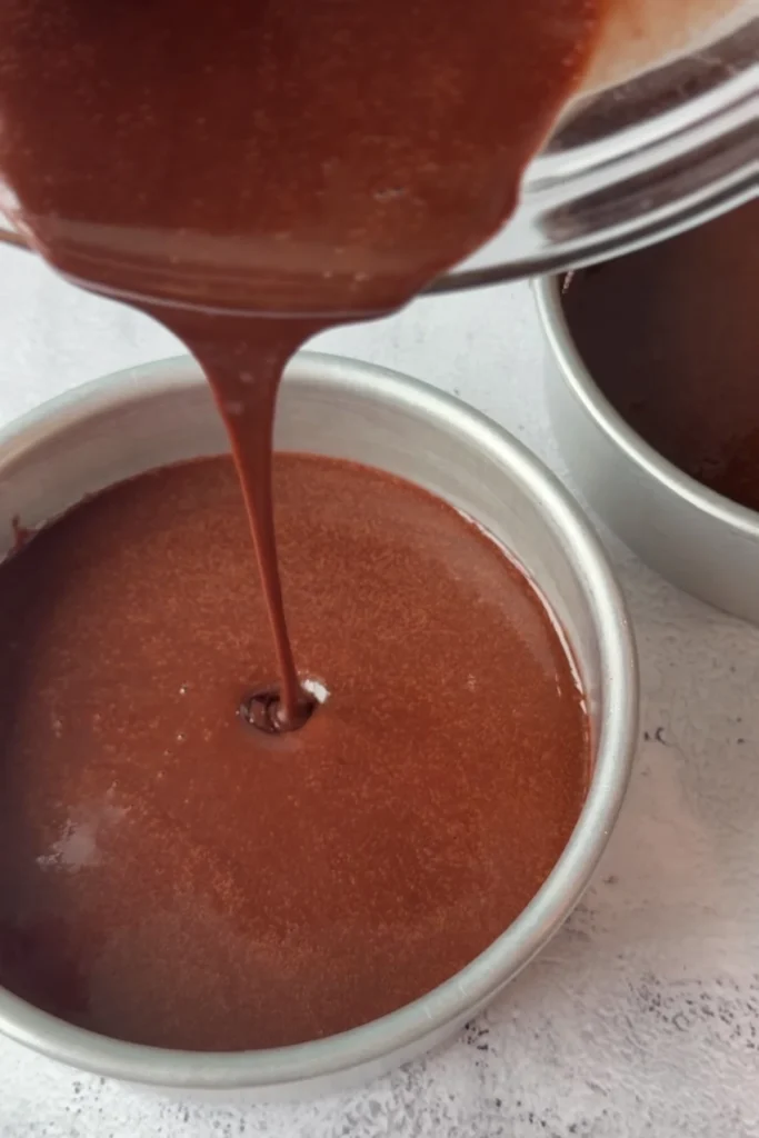 Chocolate batter being poured into a greased round cake pan, filling the bottom evenly.
