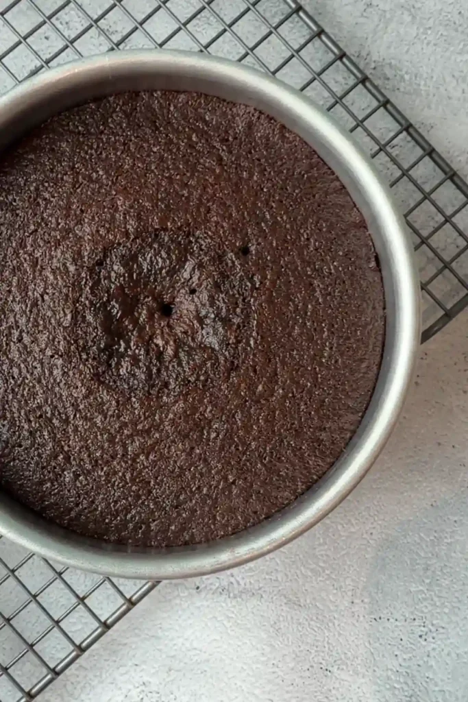 Freshly baked chocolate cake in a round pan resting on a wire rack, with a slightly domed top and a few small cracks in the center.
