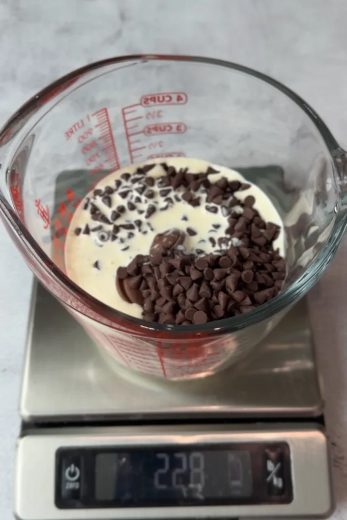Chocolate chips and cream measured in a glass cup placed on a kitchen scale, ready to be melted into ganache.