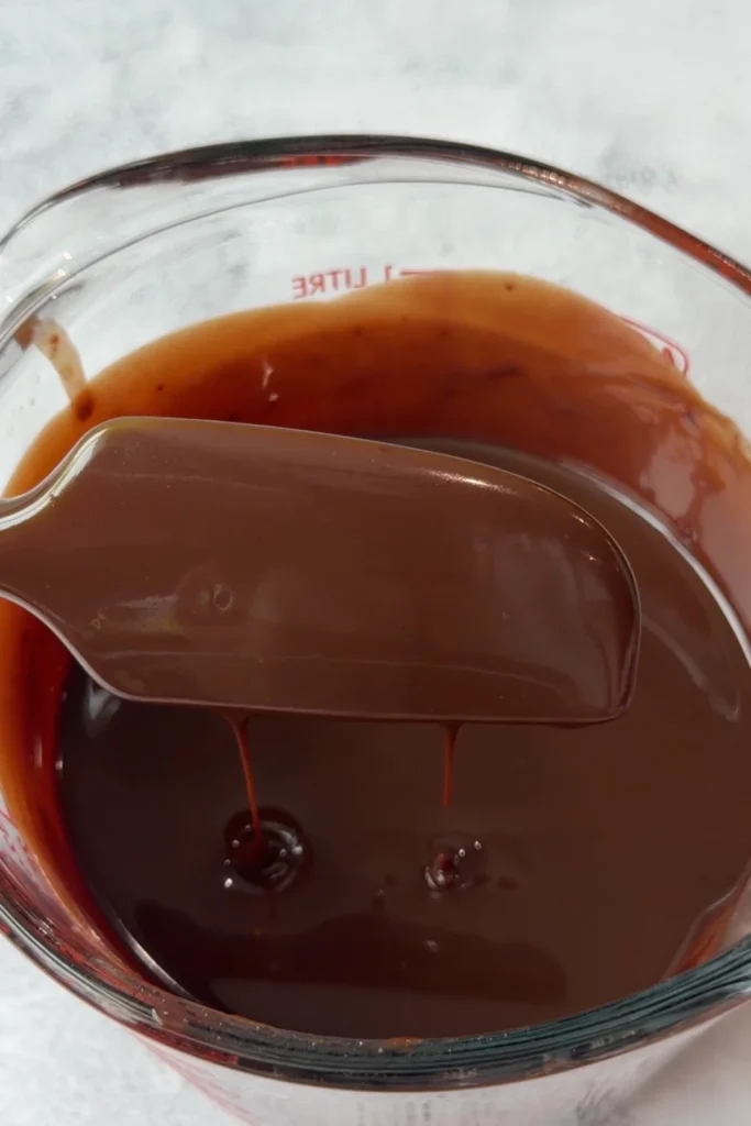 Smooth, melted chocolate ganache in a glass measuring cup, thick and glossy with a spatula lifting and dripping the mixture back into the bowl.