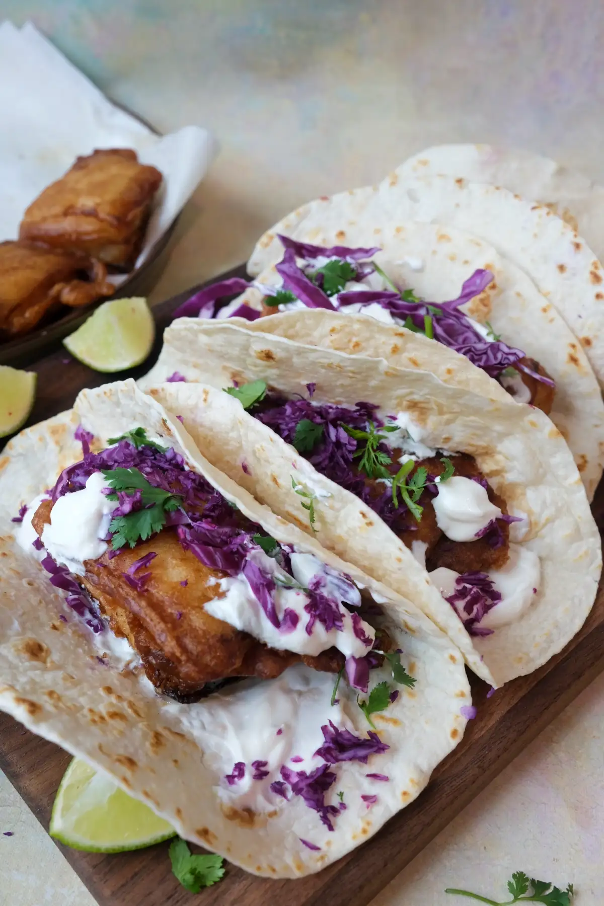 Three fish tacos on a wooden board with golden fried cod, cabbage slaw, lime wedges, and creamy sauce.