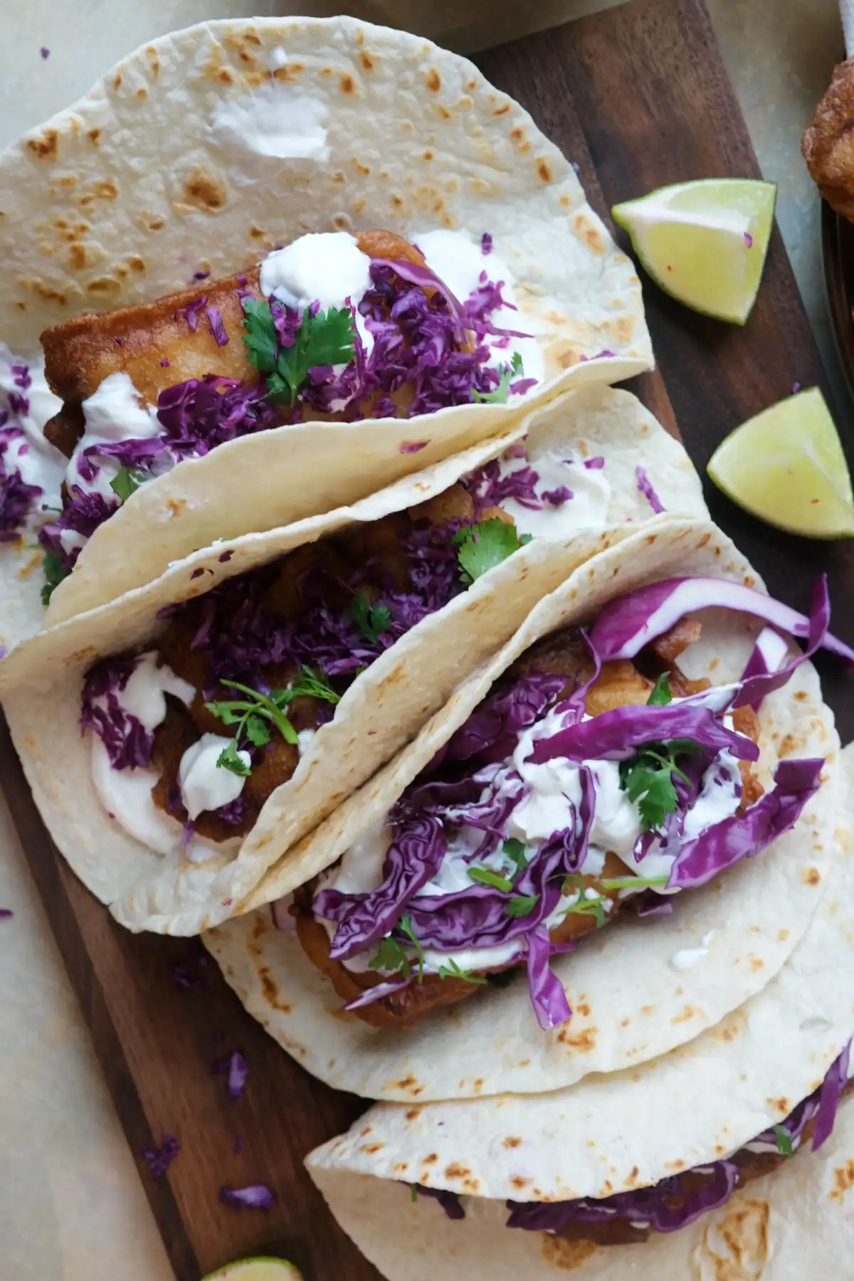 Three assembled cod fish tacos with creamy sauce, cabbage, and cilantro served on a wooden board with lime.