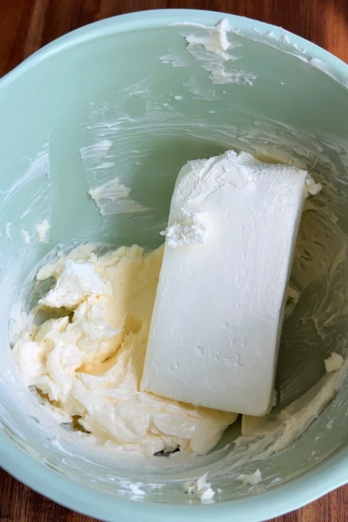 A block of cream cheese added to softened butter in a mixing bowl for the cream cheese frosting.