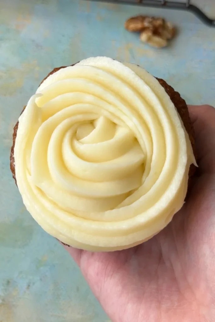 A carrot cupcake held in hand and topped with a thick spiral of cream cheese frosting piped in a rose-like swirl.