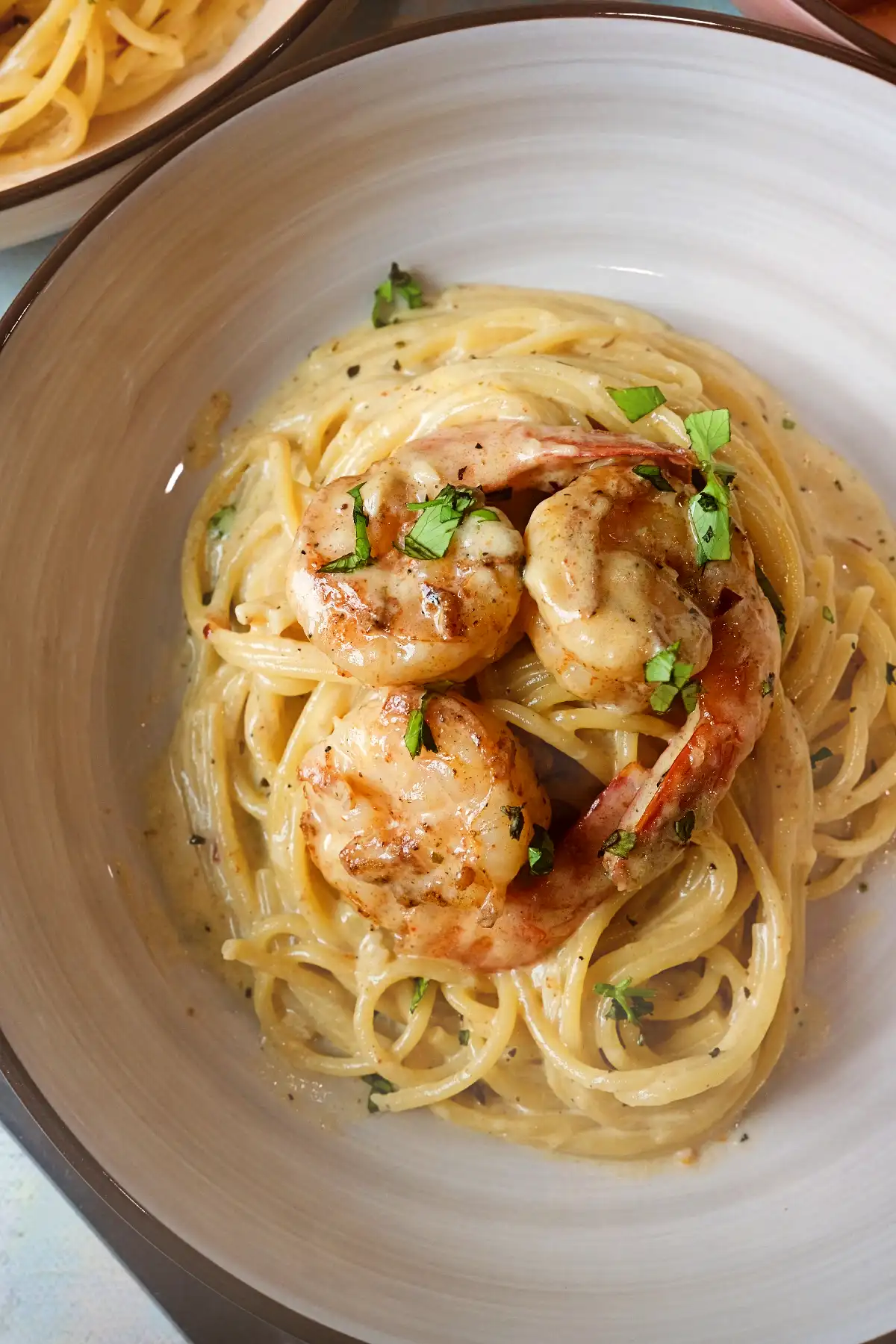 Overhead view of creamy garlic shrimp pasta in a wide beige bowl, with spaghetti coated in a silky cream sauce and topped with three seared shrimp and chopped parsley, plus a second bowl partially visible in the background.