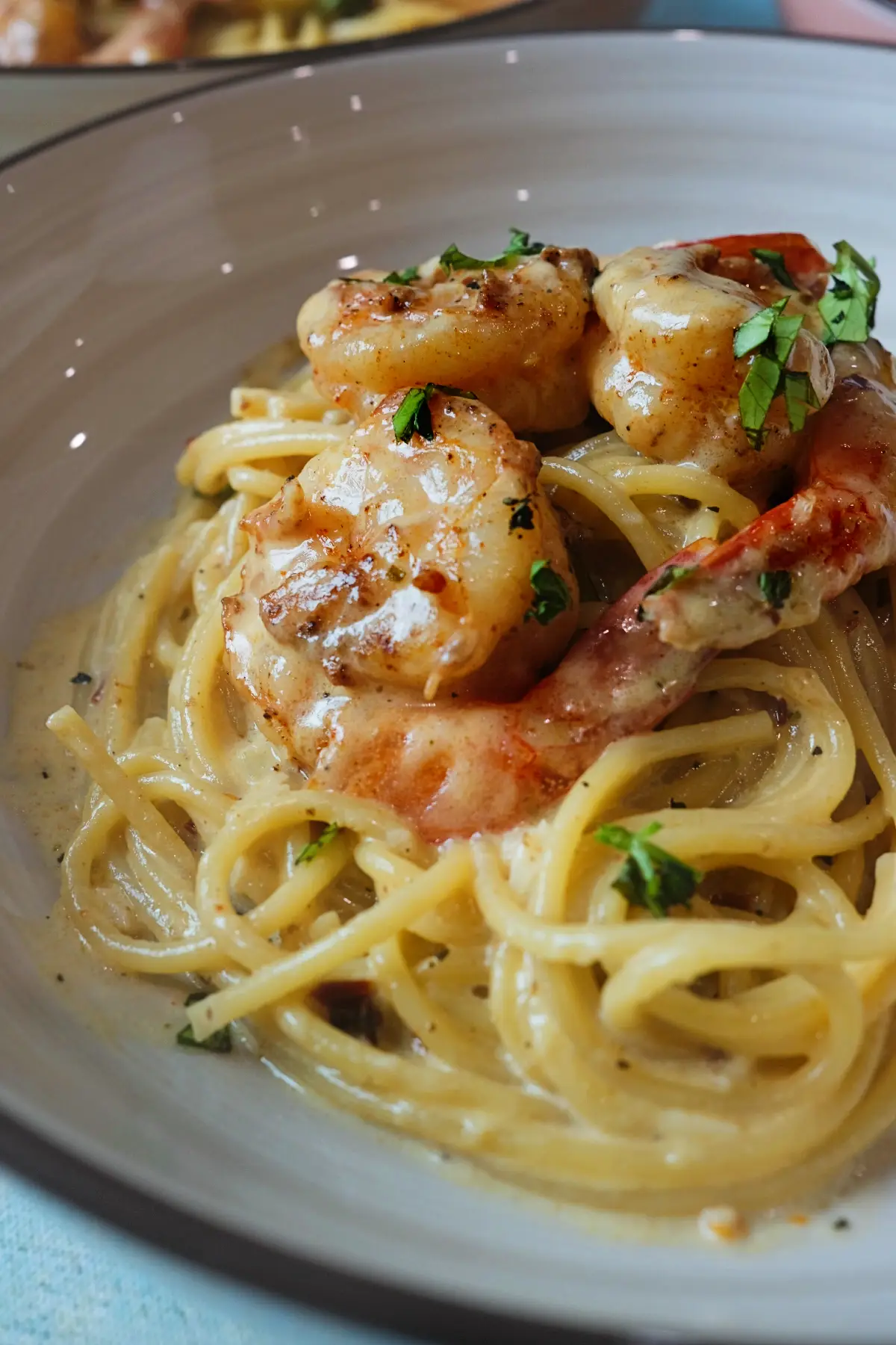 Close-up of creamy shrimp pasta in a shallow beige bowl, showing glossy spaghetti in a rich cream sauce with browned shrimp on top and scattered fresh parsley, with another pasta bowl softly blurred behind it.