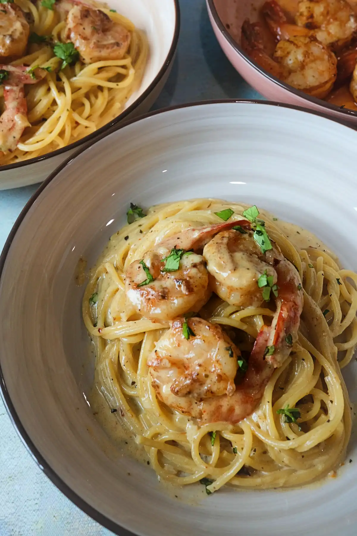 Creamy garlic shrimp pasta in a beige bowl, topped with shrimp and parsley, with a second pasta bowl and a pink bowl of extra shrimp blurred in the background.
