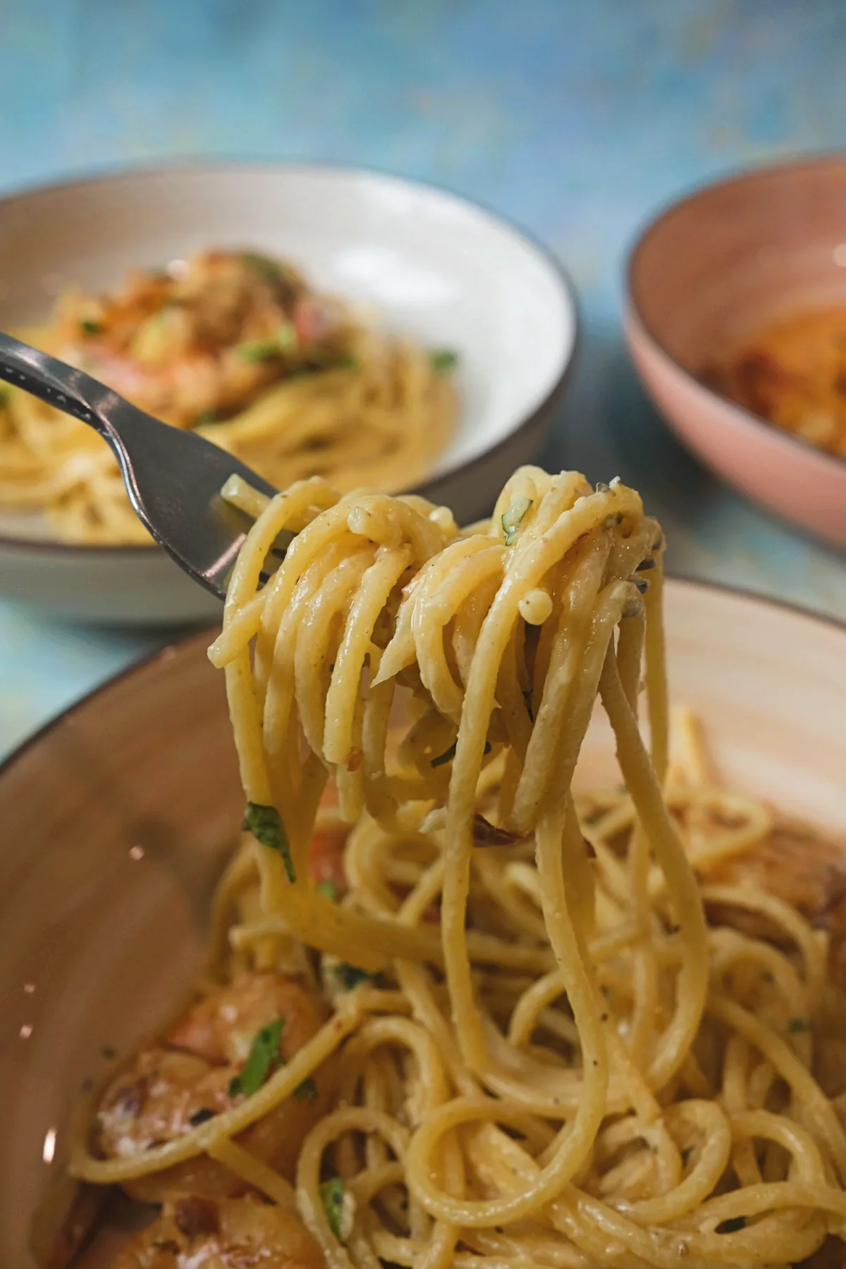 Fork lifting a twirl of creamy spaghetti above a bowl of garlic shrimp pasta, showing the smooth sauce coating the noodles, with blurred serving bowls in the background.