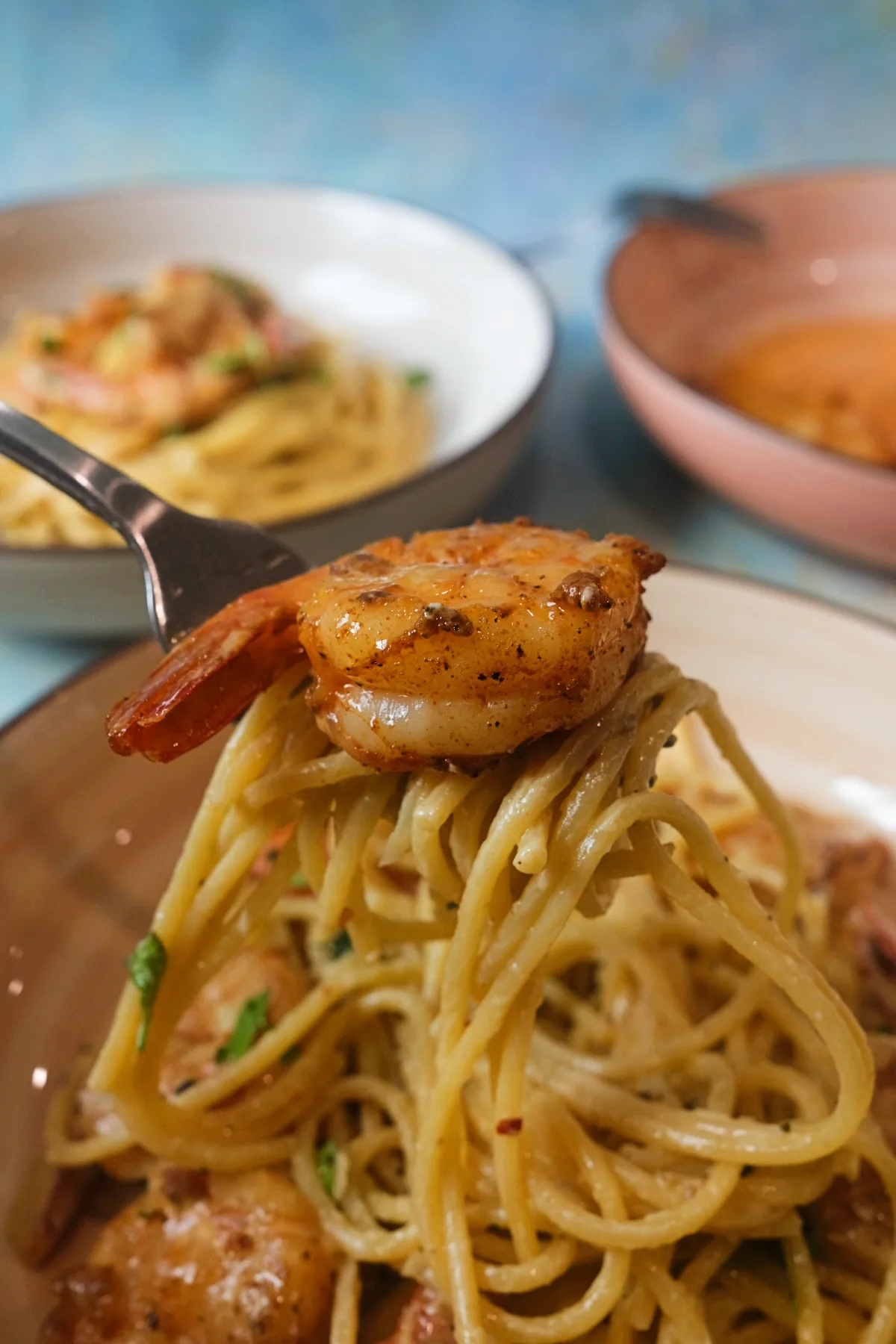 Fork holding a bundle of creamy spaghetti topped with one browned shrimp, lifted above the pasta bowl, with soft-focus bowls behind it and the sauce lightly clinging to the noodles.