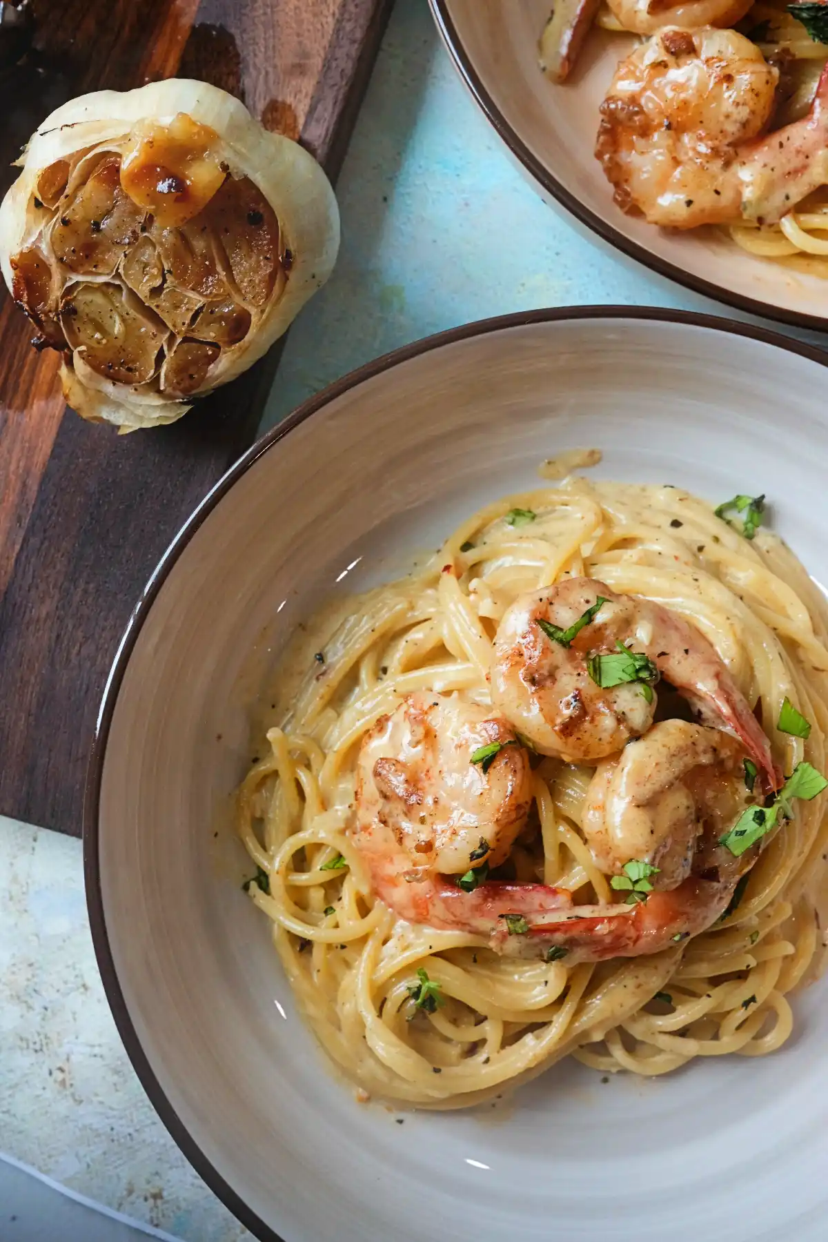 Overhead serving shot of creamy garlic shrimp pasta in a beige bowl, garnished with parsley and surrounded by a roasted garlic head on a wooden board and another bowl of pasta in the background.