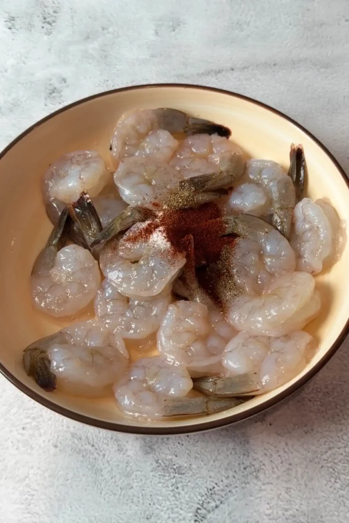 Raw peeled shrimp with tails on in a tan bowl, topped with a small pile of seasonings including salt, black pepper, and smoked paprika, set on a light stone surface before mixing.