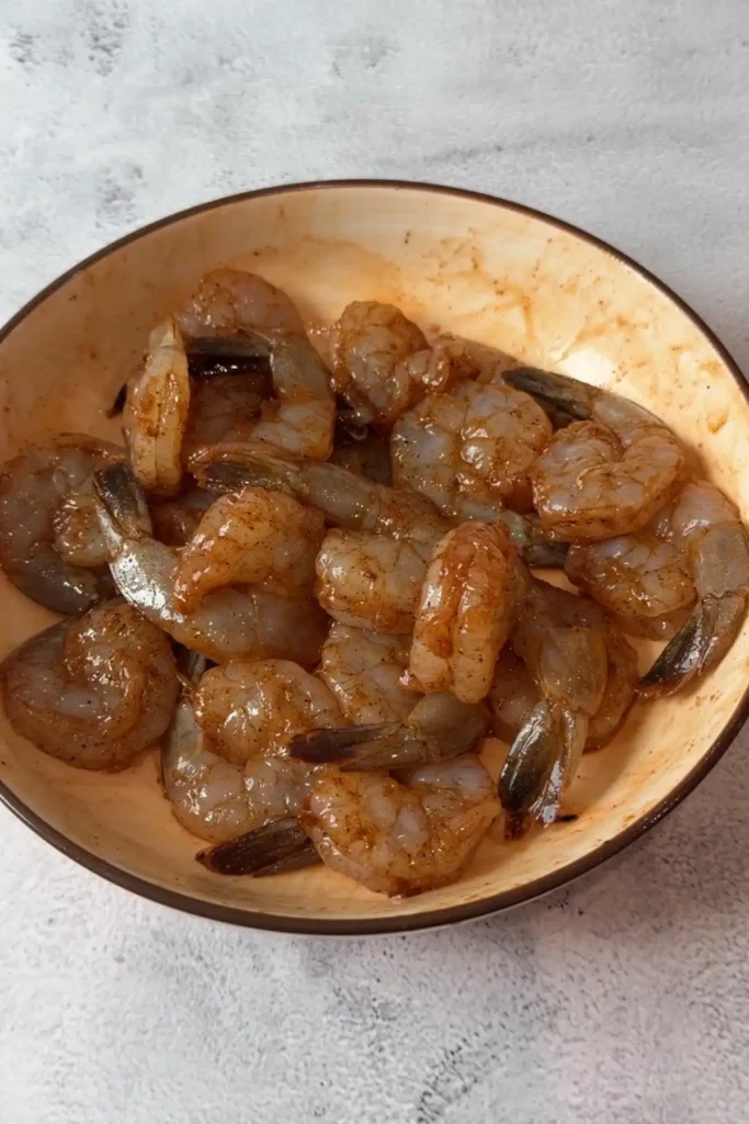 Raw shrimp in a tan bowl after being tossed with seasonings, lightly coated in a reddish paprika mixture and ready to cook, on a pale textured countertop.